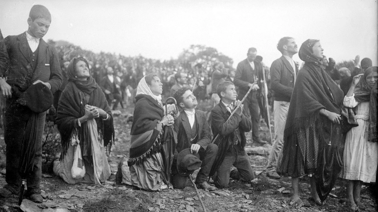 Crowd of Portuguese villagers in shawls and caps kneeling and standing on rocky ground at Fatima on 13 October 1917, all of them staring upward at the sky