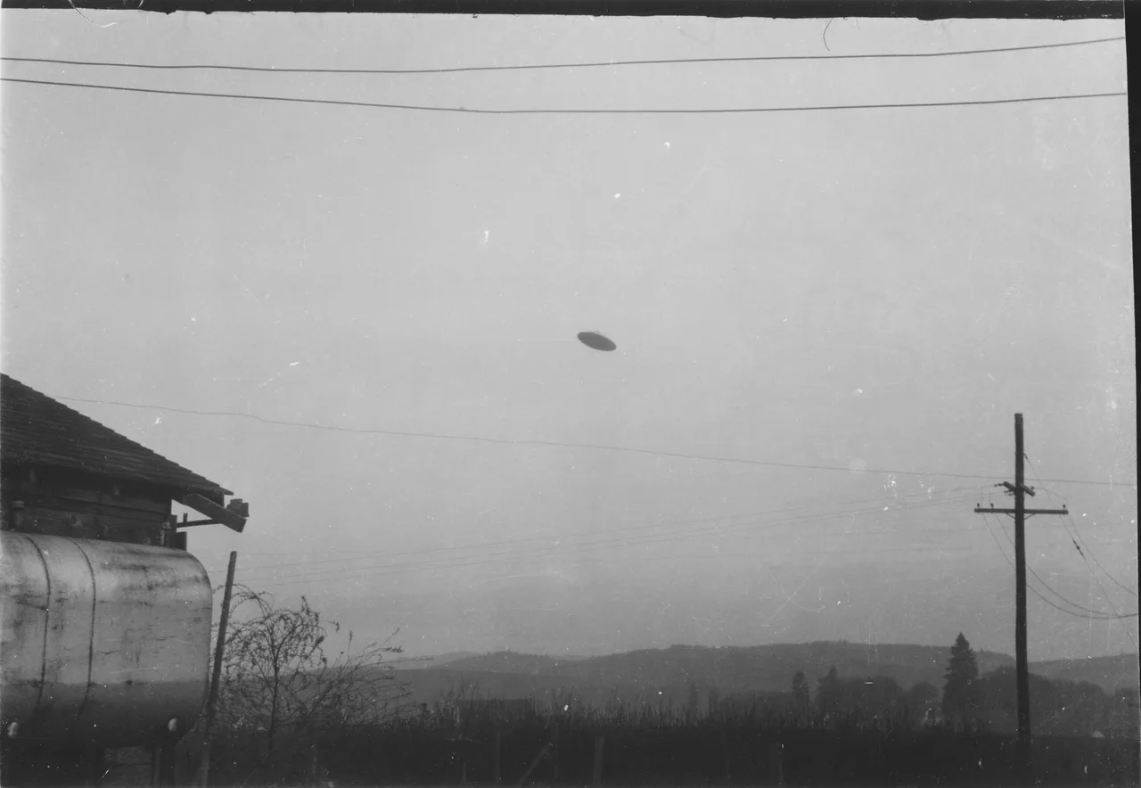 Black and white photograph of a small flat disc-shaped object in a grey sky above a farm, with power lines across the top of the frame and a utility pole at right