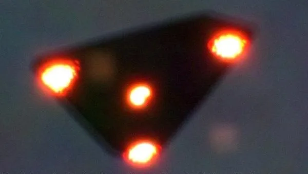Blurry photograph of a dark triangle against a grey-blue sky with a bright orange light at each corner and one in the centre