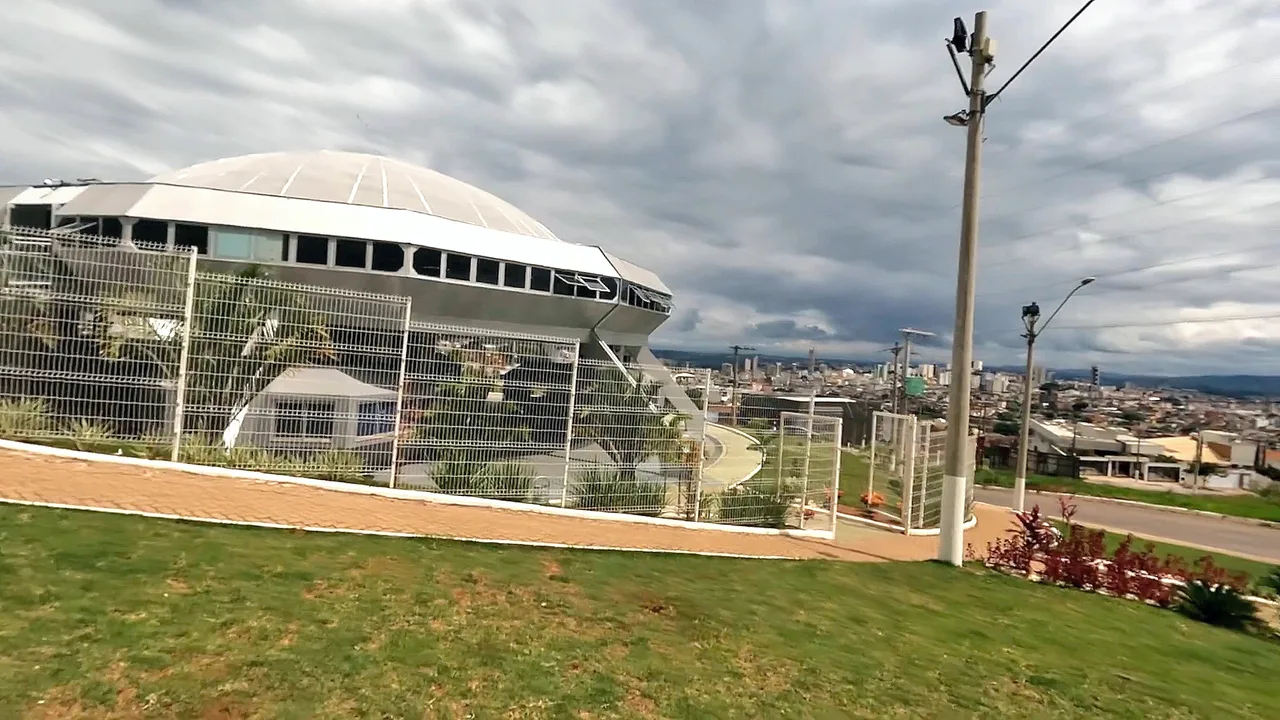 A white flying-saucer-shaped building on a hill above a Brazilian city under a cloudy sky, behind a wire fence