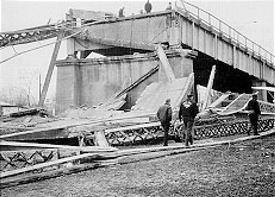 Black and white 1967 photograph of the collapsed Silver Bridge from the Ohio bank, the deck buckled into the water with men standing among the wreckage
